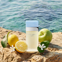 Perfume bottle with lemons and an apple on a rocky surface by water